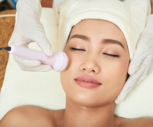 Close-up shot of beautician hands making ultrasound facial procedure while pretty Asian client lying on treatment table with closed eyes